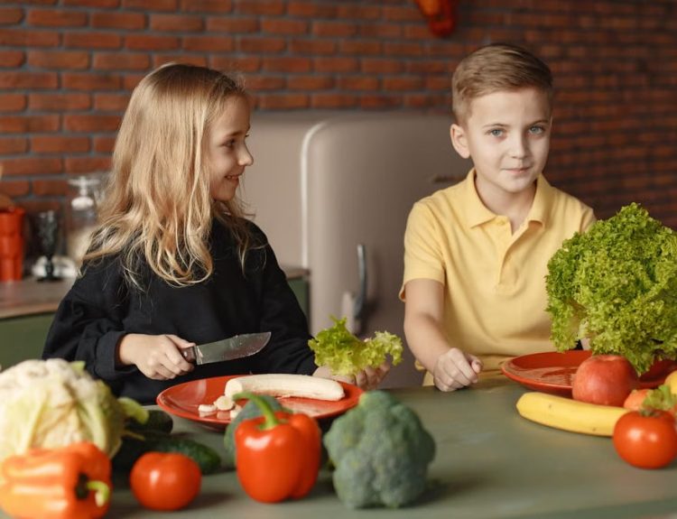 Positive little girl and boy sitting at table with assorted vegetables and fruits while preparing healthy food together in loft kitchen with brick wall